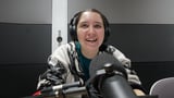 Woman wearing headphones and a patterned jacket speaks into a microphone in a recording studio