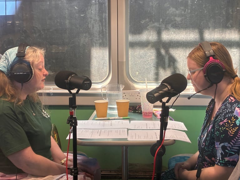 Two women recording a podcast in a bright room with microphones, headphones, and drinks on the table between them